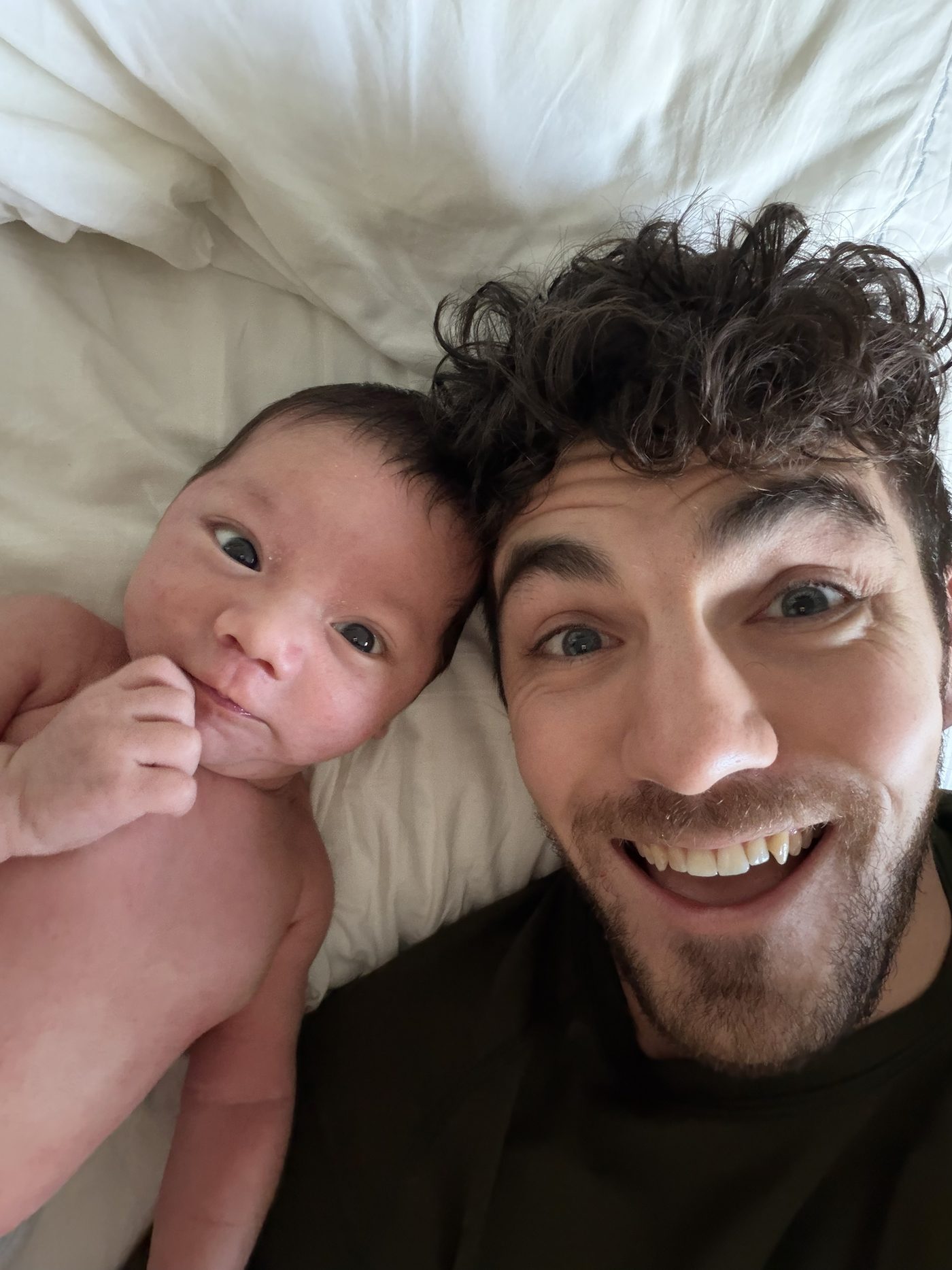 Alex and his newborn son Hudson, cheek to cheek on a white bed, both looking up at the camera